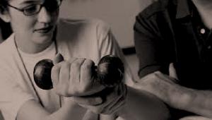 FotografÃ­a en blanco y negro de una persona realizando ejercicio fÃ­sico con una pesa en la mano supervisada por una especialista con gafas